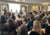 A room full of seated people listen to a speaker.